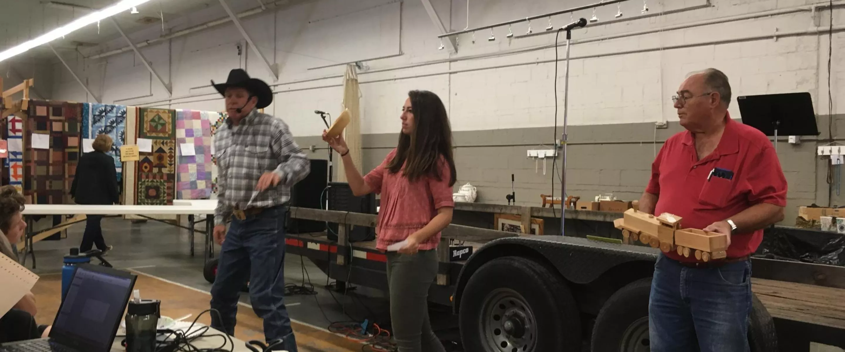 An auctioneer in a black cow boy hats stands next to a woman holding auction item, a wooden bowl. Next to here is a man holding another auction item, a wooden toy train.