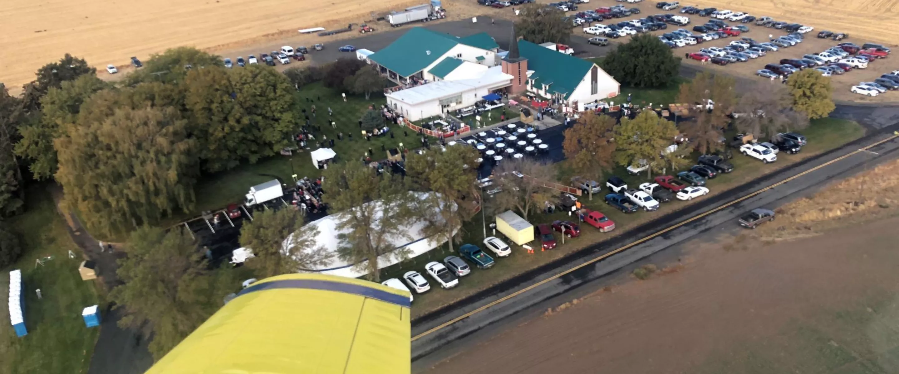 An overview view of a church building during an event. There are a lot of cars parked around the church. The photo was taken out the window of a yellow airplane.