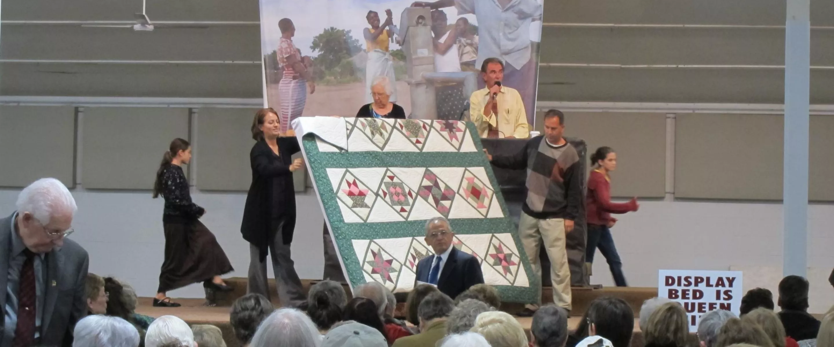 Three people display a quilt during an auction. Behind them is an auctioneer.