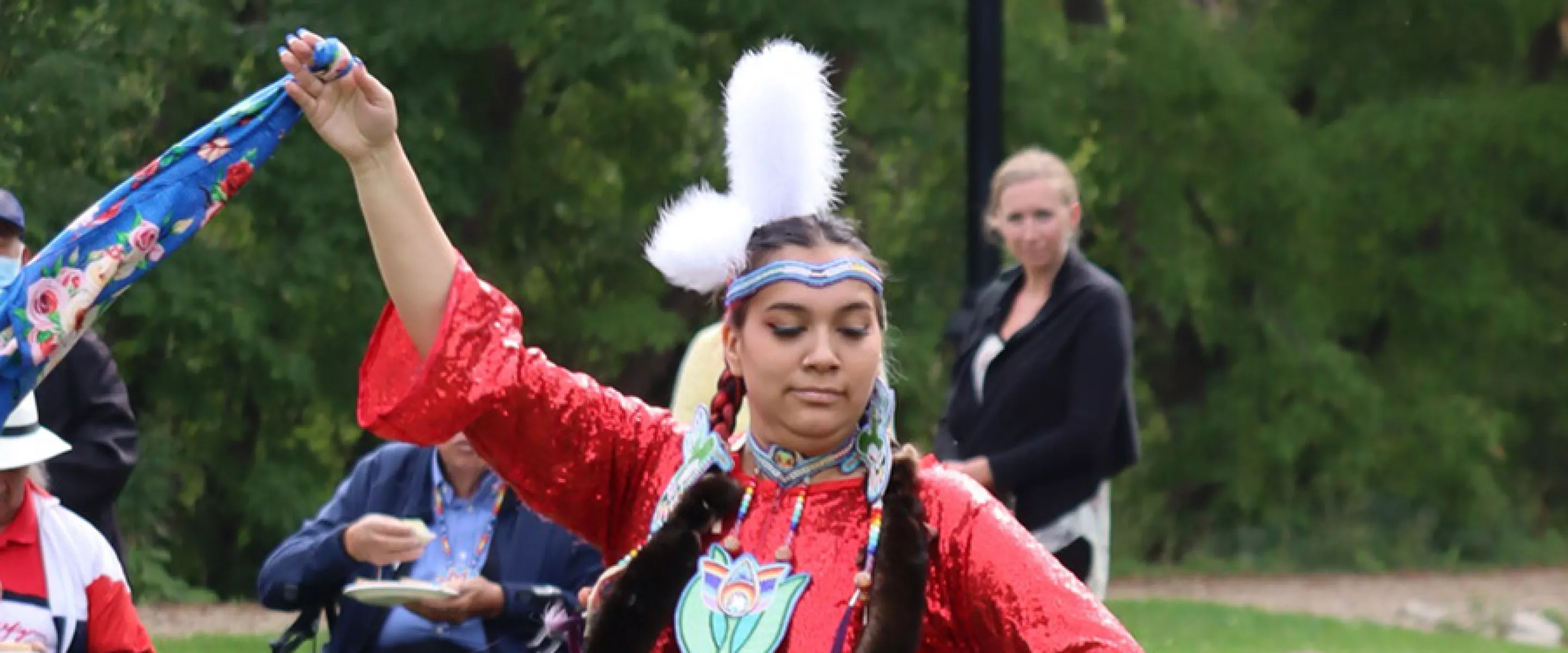 An Indigenous woman in traditional clothing dances and waves a scarf in the air while onlookers watch.