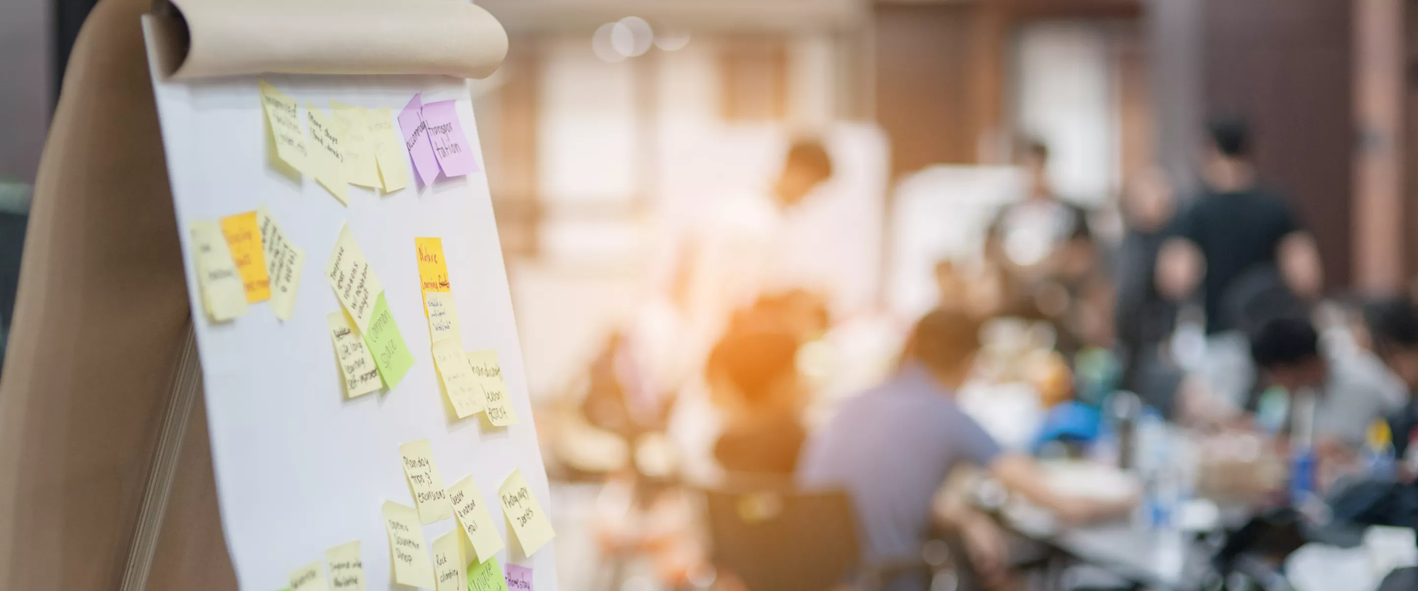 A flip chart board is in the foreground with sticky notes attached, while a group sits in the background.