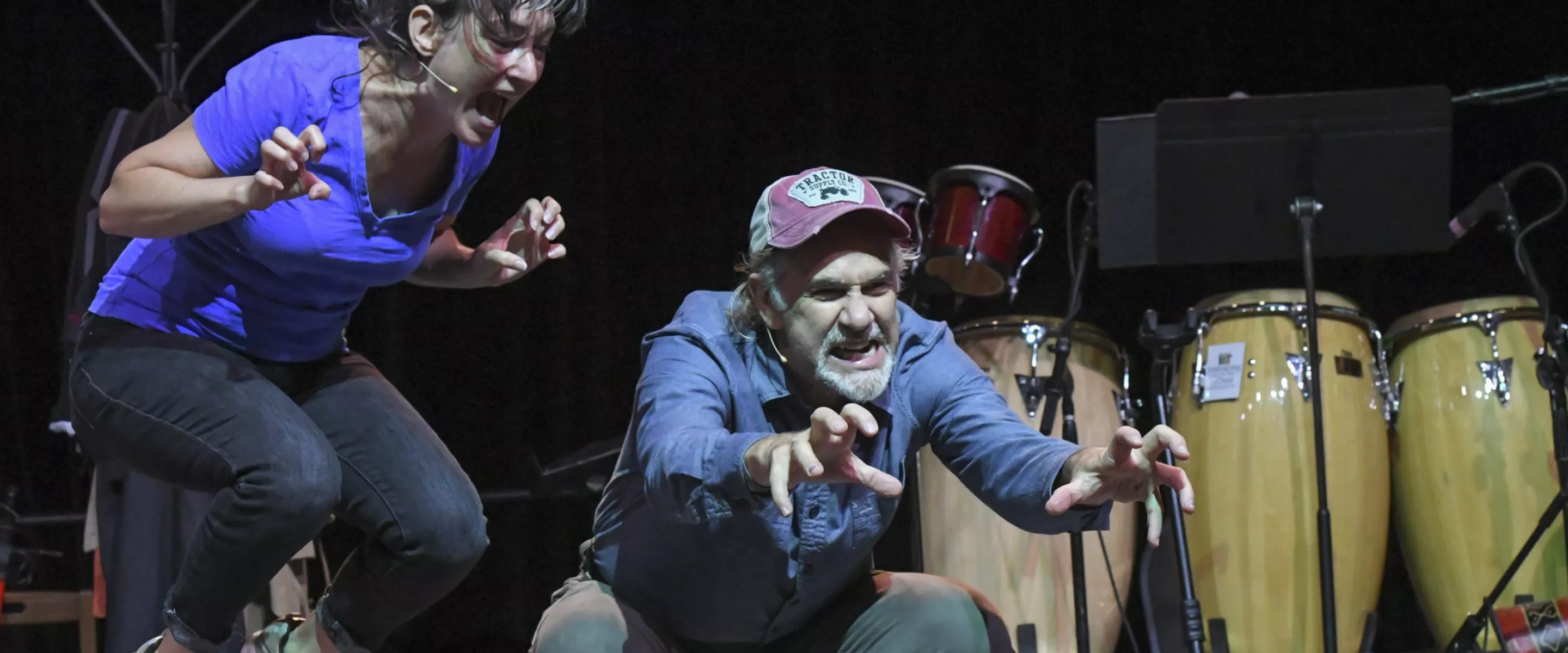 A man and a woman act on stage. The woman is jumping in the air, and the man is crouching.