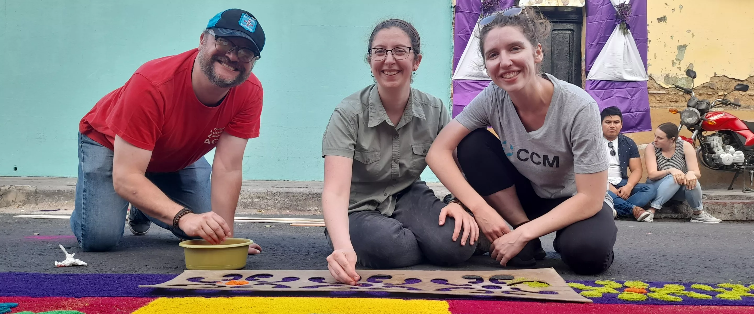 Two women and a man are crouching and smiling at the camera in front of a colourful canvas.