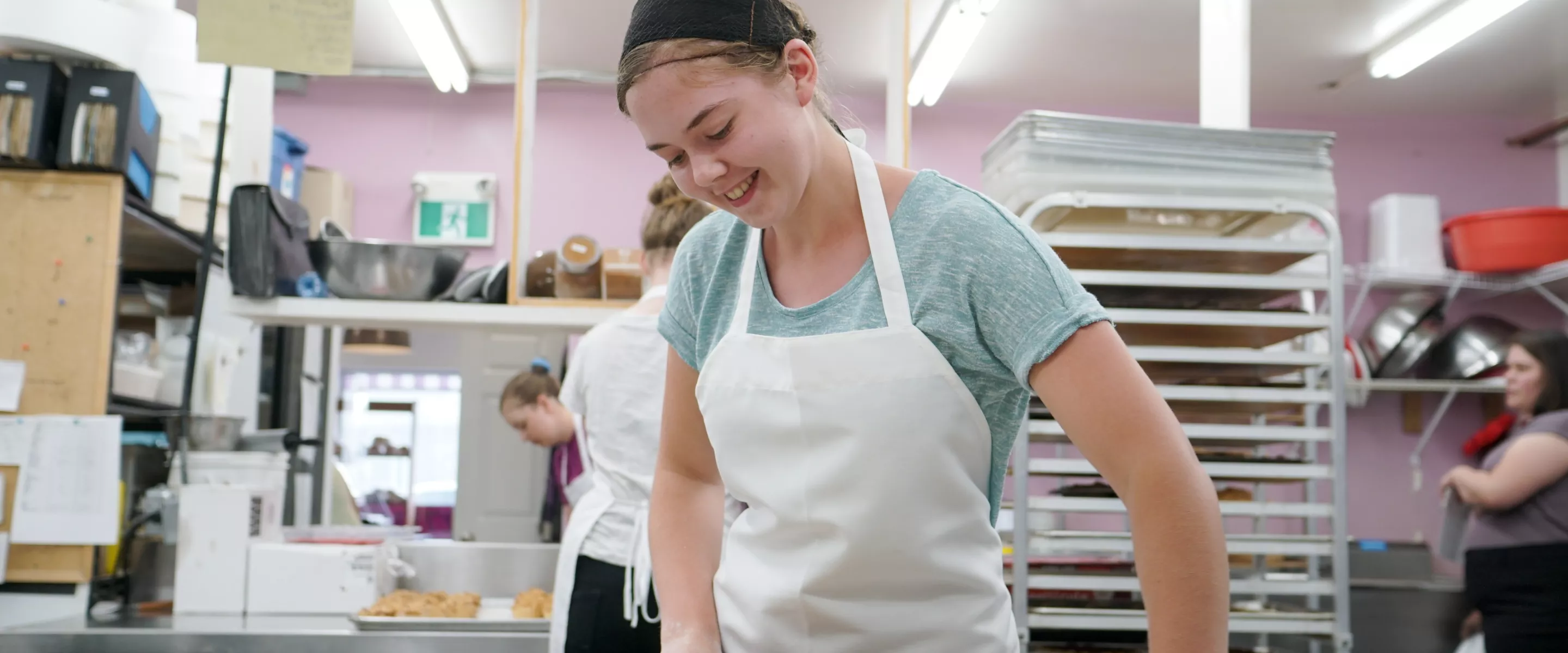 A young lady with a white apron uses a rolling pin to roll out dough.