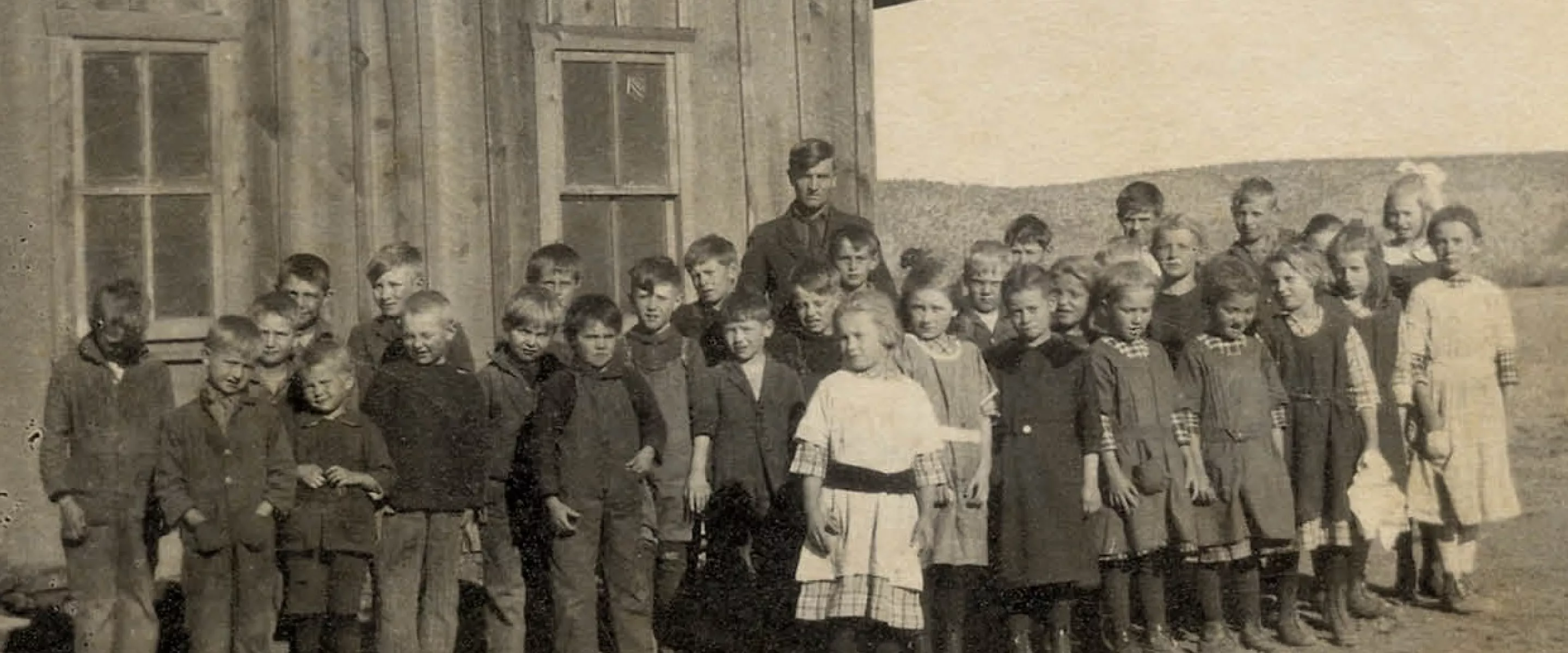 An old photograph of several school children standing in front of a wooden building with one adult among them.