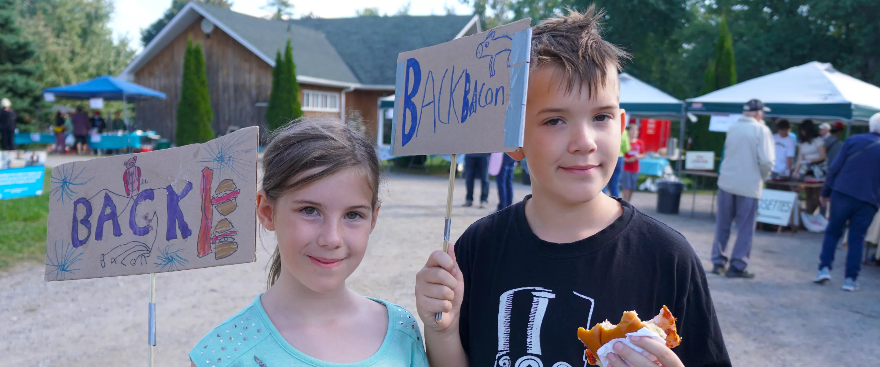 A boy and a girl hold up hand made cardboard signs that read Back Bacon, while the boy eats bacon on a bun.