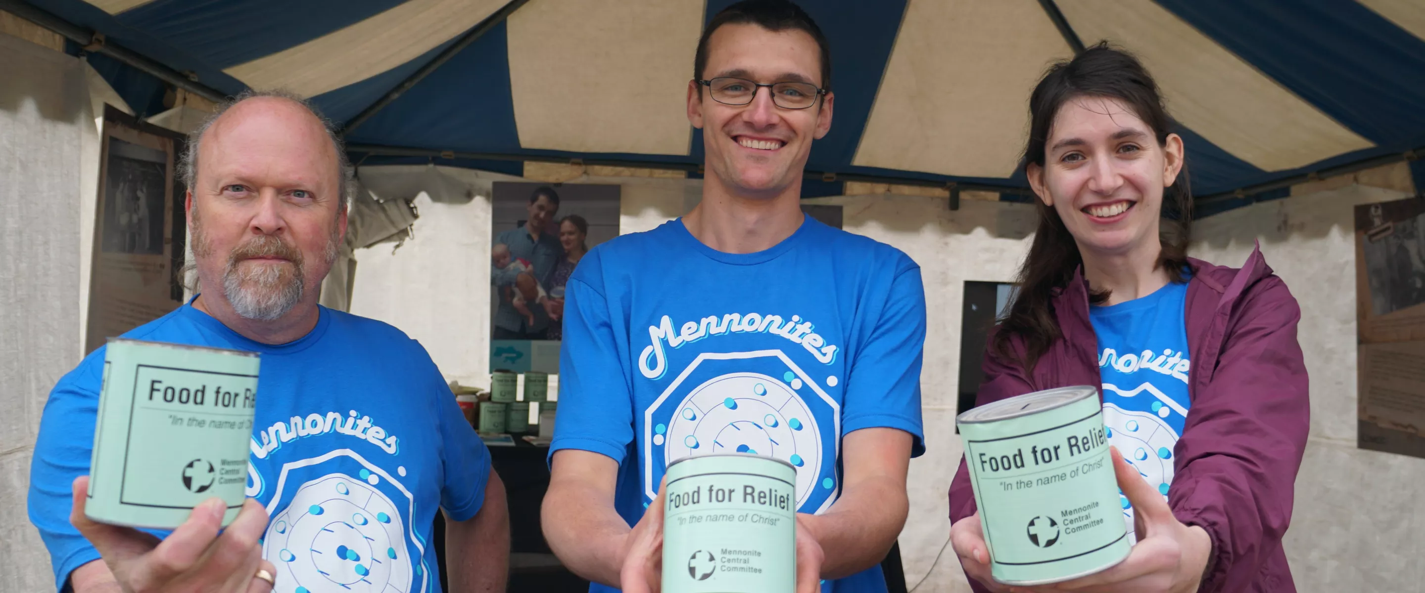 Standing outside a tent, three people wearing identical blue shirts each hold out a can that reads "Food for Relief" to the camera.