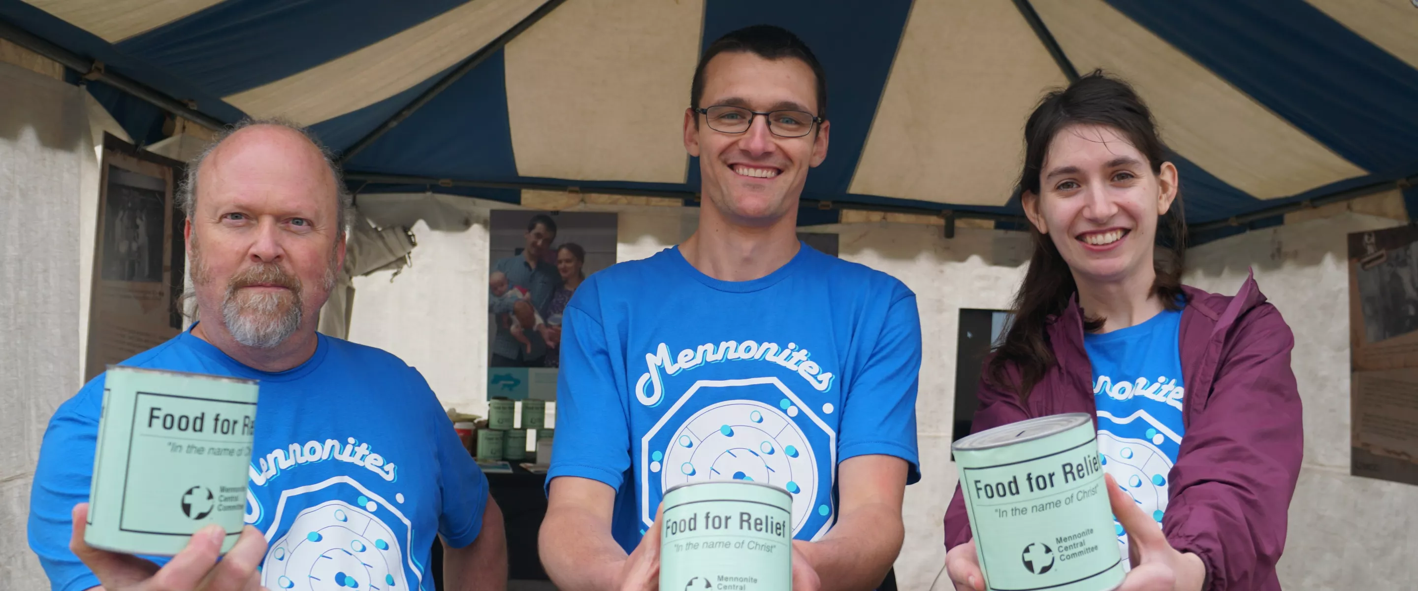 Standing outside a tent, three people wearing identical blue shirts each hold out a can that reads "Food for Relief" to the camera.