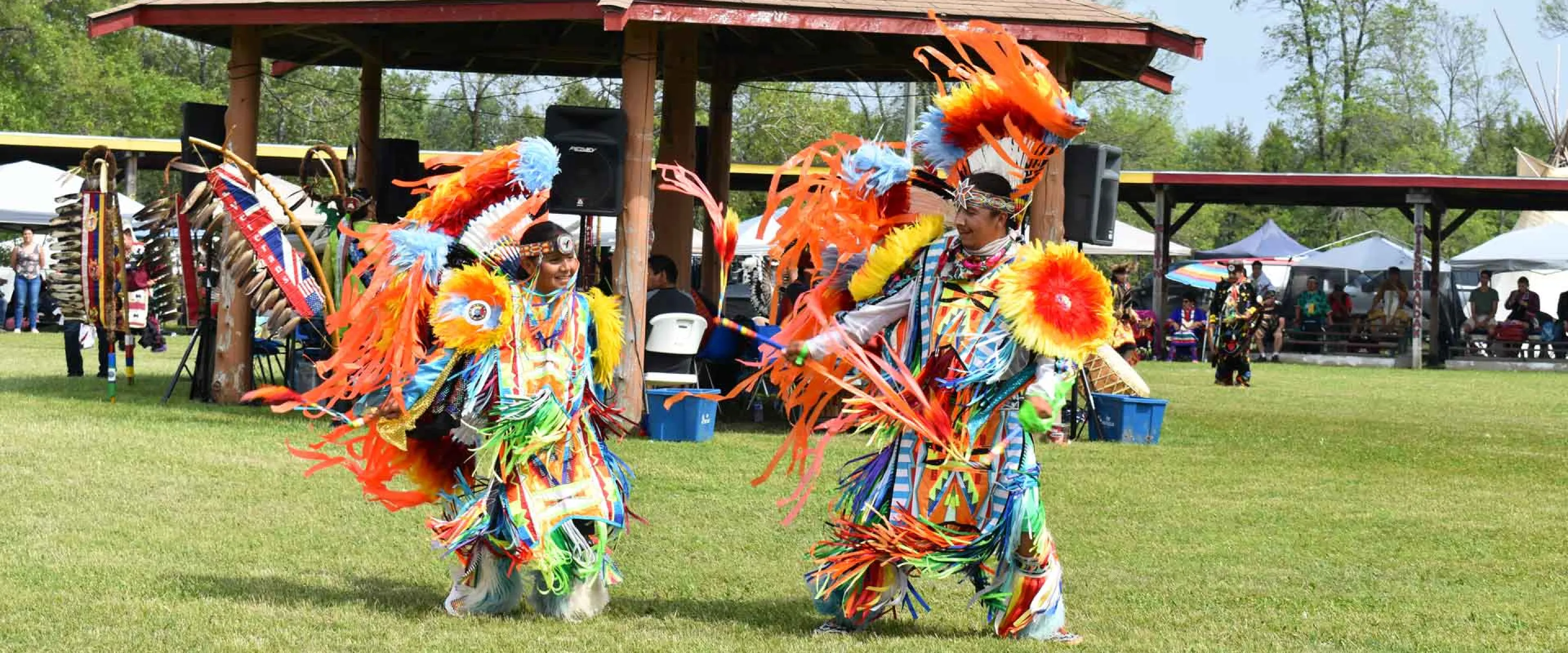 A father and son dance in colourful traditional Indigenous dress in front of a pavillion on green grass.