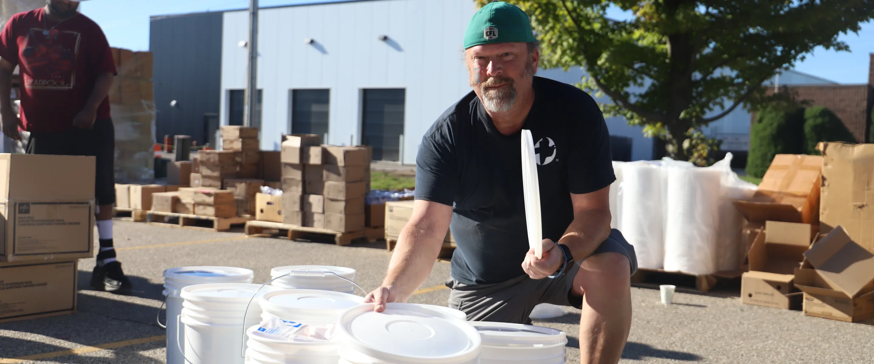 A man with a cap is kneeling next to several white buckets. Boxes are in the background.