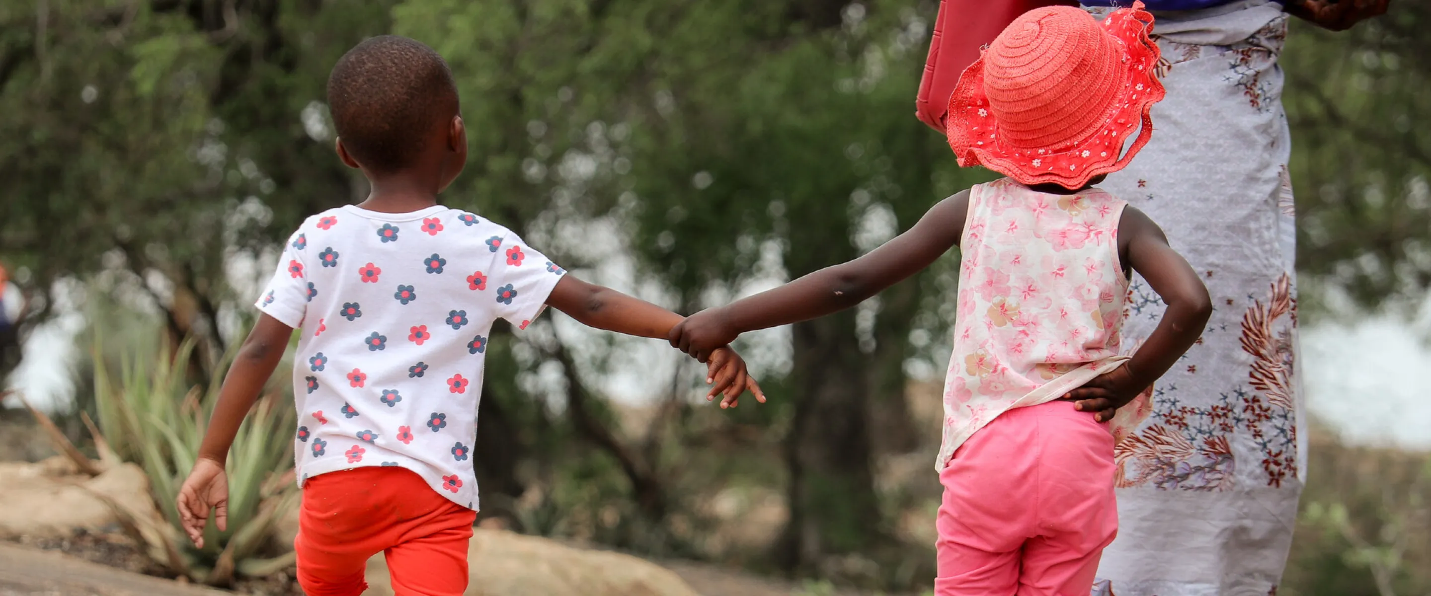 Two children in Zimbabwe hold hands while walking down the road. One is wearing a bright coloured hat.