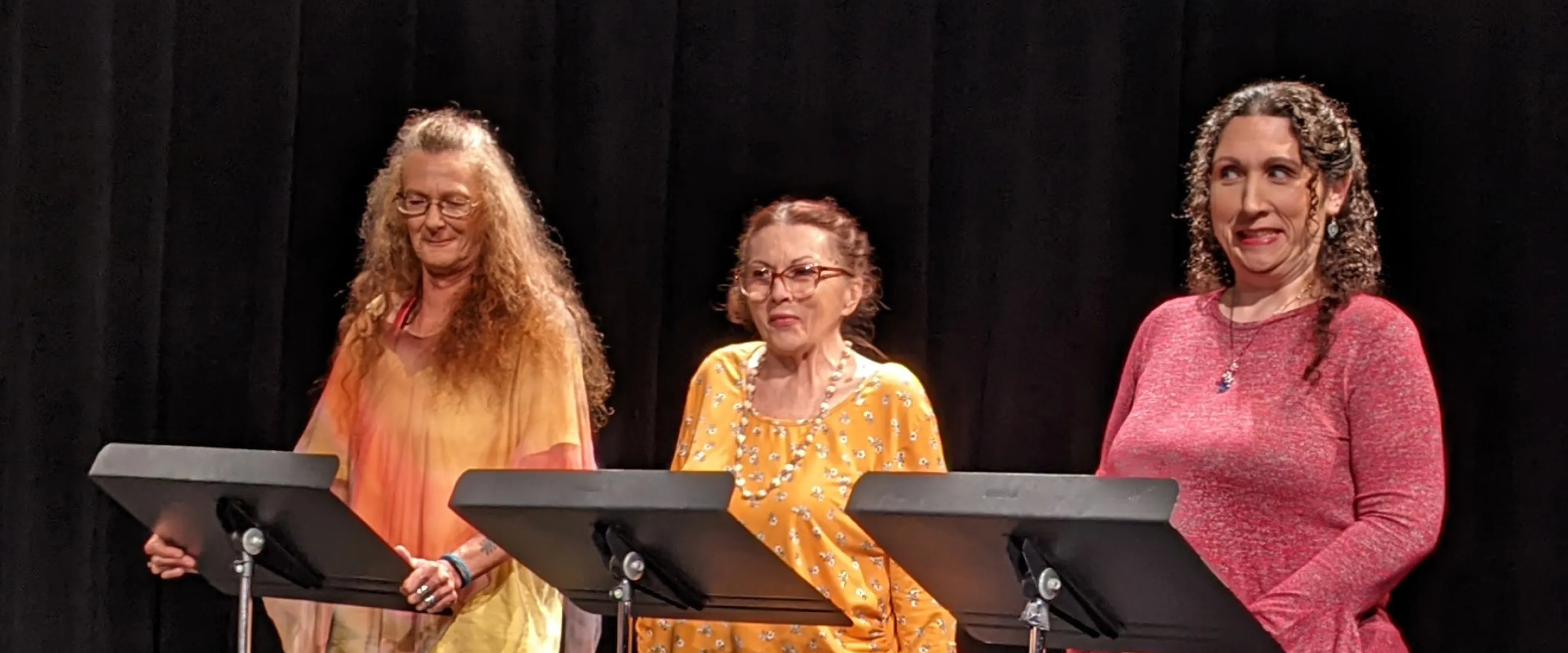 Three women stand behind music stands in a dark room.