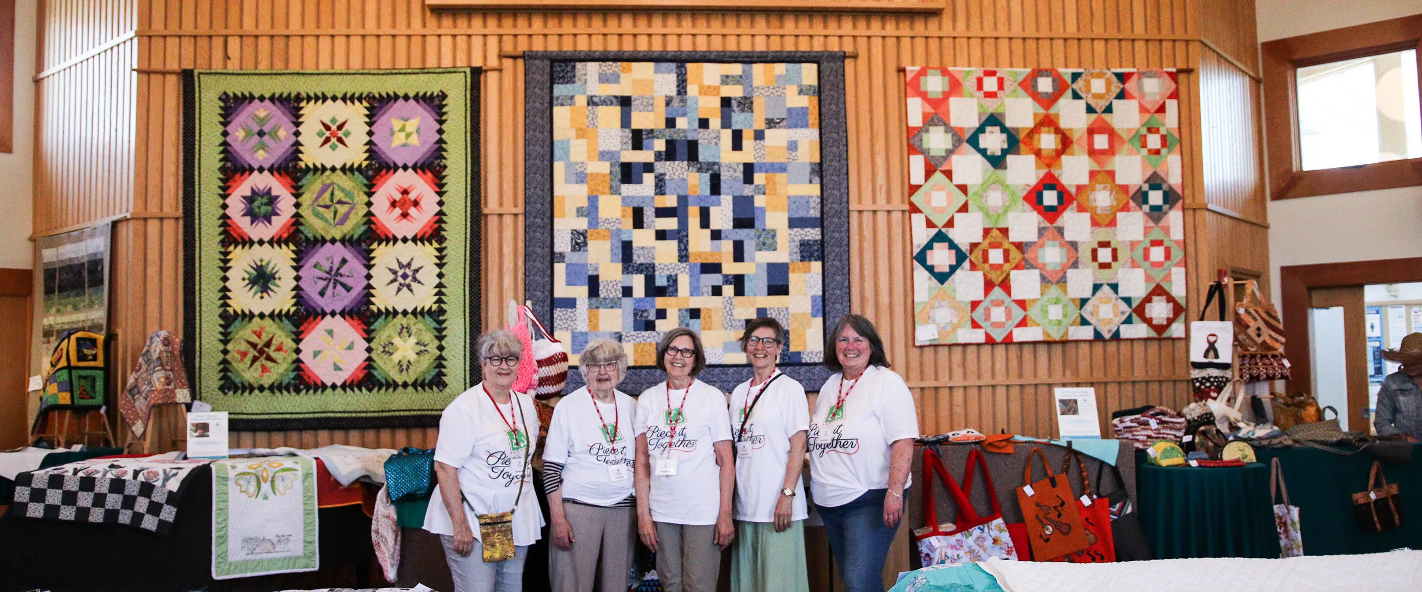 Five members of the Piece It Together quilting group stand in front of three quilts during an annual sale