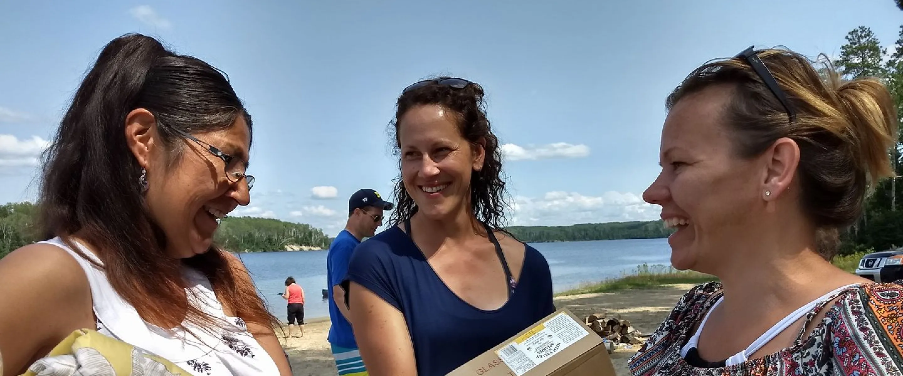 Three women holding boxes and comforters while standing near a shoreline.