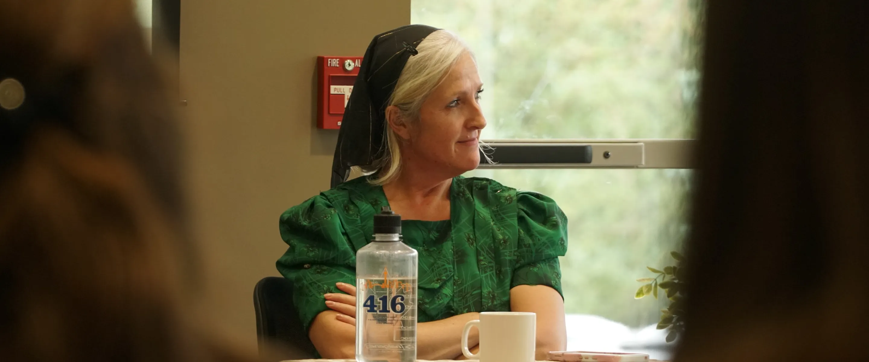 A woman dressed in traditional Mennonite clothing and head cover sits at a table in conversation.