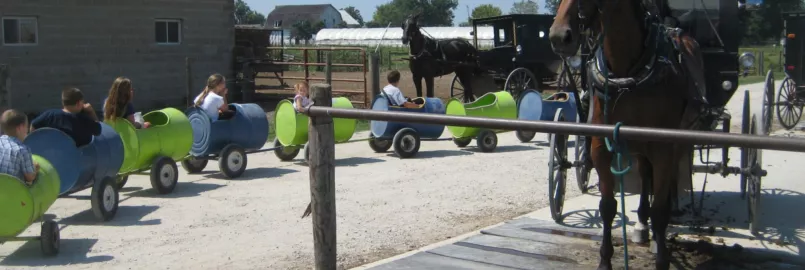 Children ride in barrels on wheels. There are Amish buggies and horses parked nearby.