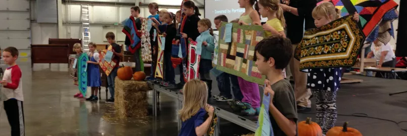 Several children stand on a stage holding up quilted wall hangings. There is an older woman behind them speaking into a microphone. 