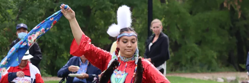 An Indigenous woman in traditional clothing dances and waves a scarf in the air while onlookers watch.