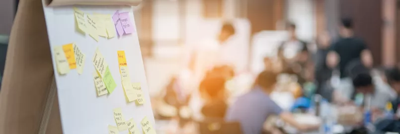 A flip chart board is in the foreground with sticky notes attached, while a group sits in the background.