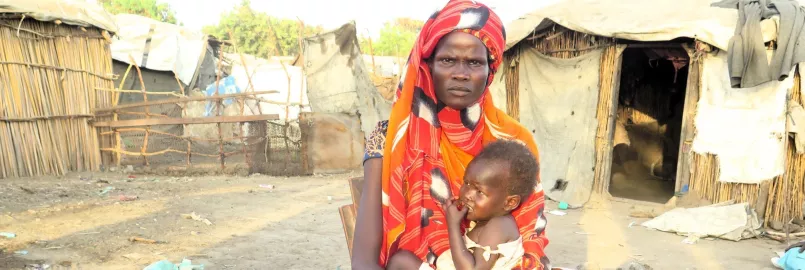A woman holding a baby in front of a camp for internally displaced people.