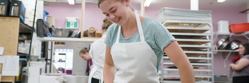 A young lady with a white apron uses a rolling pin to roll out dough.