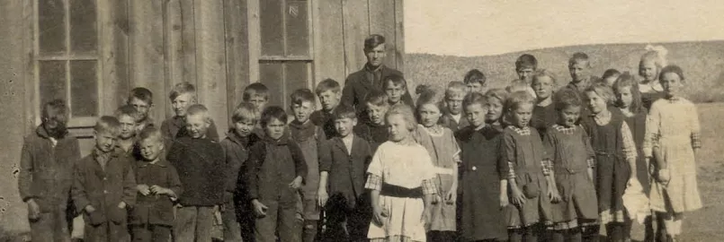 An old photograph of several school children standing in front of a wooden building with one adult among them.