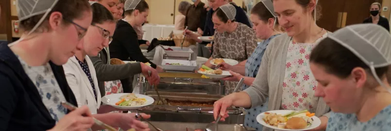 Ladies with traditional dress and head coverings gather around both sides of a buffet table.