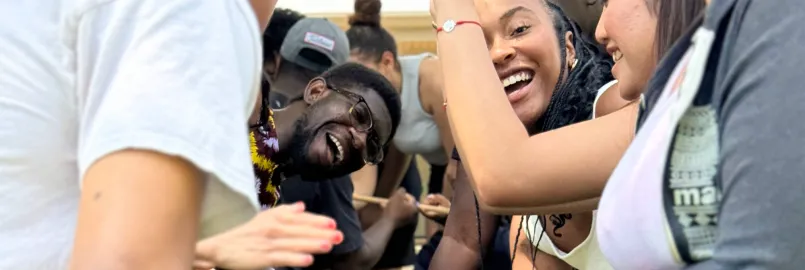 A group of smiling young people participate in a team-building activity that involves a stick on the ground.