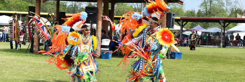 A father and son dance in colourful traditional Indigenous dress in front of a pavillion on green grass.