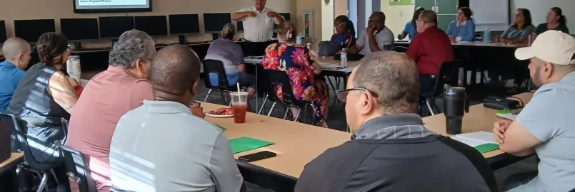 group of people sitting around a few tables listening to the main facilitator standing in the center