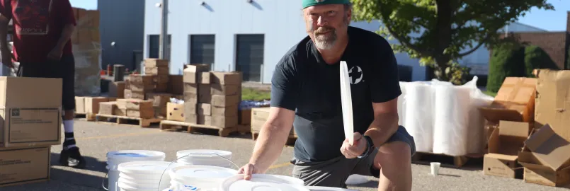 A man with a cap is kneeling next to several white buckets. Boxes are in the background.