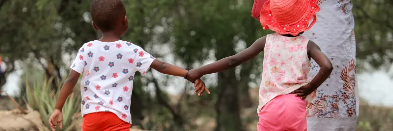 Two children in Zimbabwe hold hands while walking down the road. One is wearing a bright coloured hat.