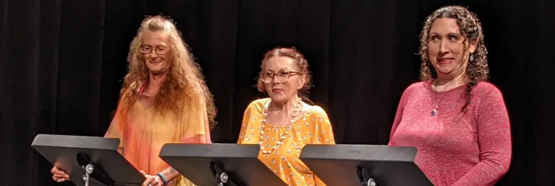 Three women stand behind music stands in a dark room.