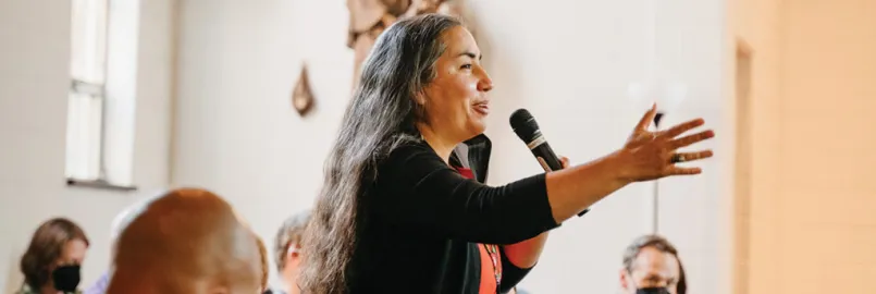 Sarah Augustine stands and holds a microphone while talking to a crowd of people sitting down.