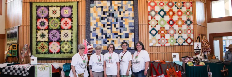 Five members of the Piece It Together quilting group stand in front of three quilts during an annual sale