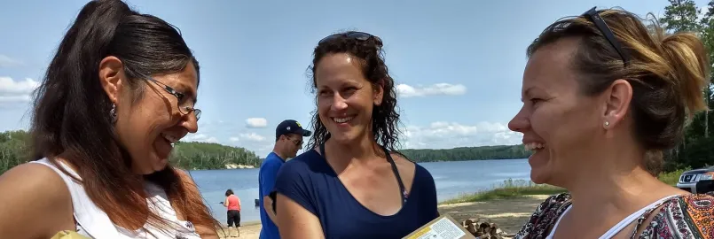 Three women holding boxes and comforters while standing near a shoreline.