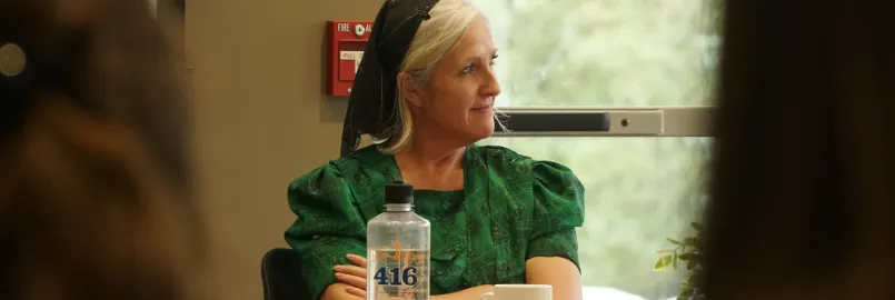 A woman dressed in traditional Mennonite clothing and head cover sits at a table in conversation.