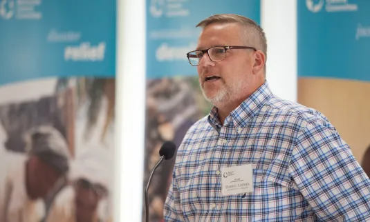 A man in a plaid shirt and glasses speaks into a microphone