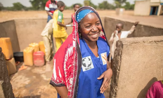 A Chadian woman in a blue shirt and red scarf covering her read laughs near a well.