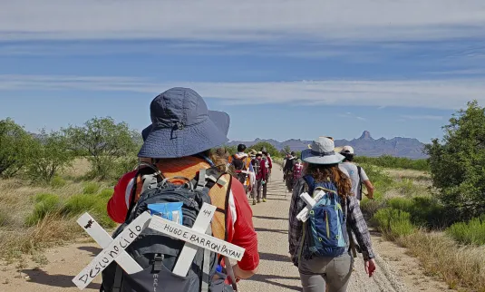 A group of people are walking on a dirt trail. There are crosses in their backpacks with messages in Spanish.