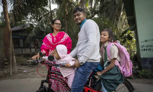 Un hombre se sienta en una bicicleta con dos niñas y una mujer cerca. Una de las niñas lleva una mochila y delante hay un bebé sentado. Al fondo se ven árboles.