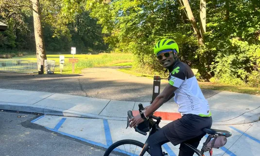 A person on a bicycle, wearing a helmet, smiles while positioned near a path surrounded by trees and greenery.