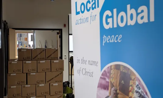 The MCC Manitoba Global/Local banner sits in front of a pallet of relief kits at the MCC Manitoba office