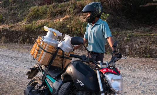 + A person wearing a helmet stands beside a motorcycle loaded with various items, including containers. The setting appears to be a rural road during daylight.