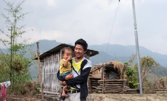 + A man smiles holding a child, standing outdoors near a wooden shack with mountains in the background under a cloudy sky. There's a sense of rural simplicity and contentment.