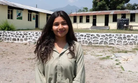 + A woman is smiling in front of a two-story building with trees and motorcycles parked nearby, under a clear sky.