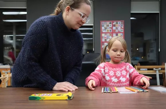 A woman leans over a child as the child counts out colored pencils on a table.