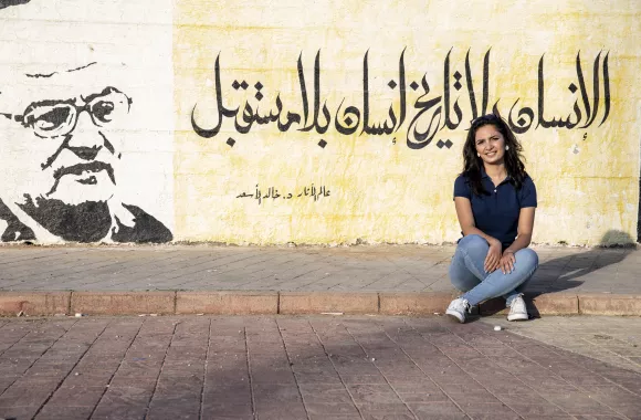 Mural project coordinator Hourya Mohammed sits in front of a wall of murals painted in the city of Tartus, Syria, in 2019. The yellow panel behind her depicts a quote from al-Asaad, “A person with no history, is a person with no future.”
