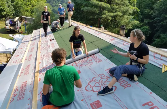 Three women sit on a roof while working on shingling it