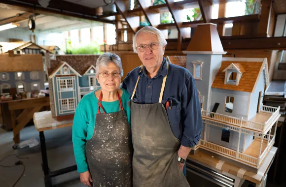 An older man and woman stand in a workshop featuring dollhouses. They are both wearing work aprons.